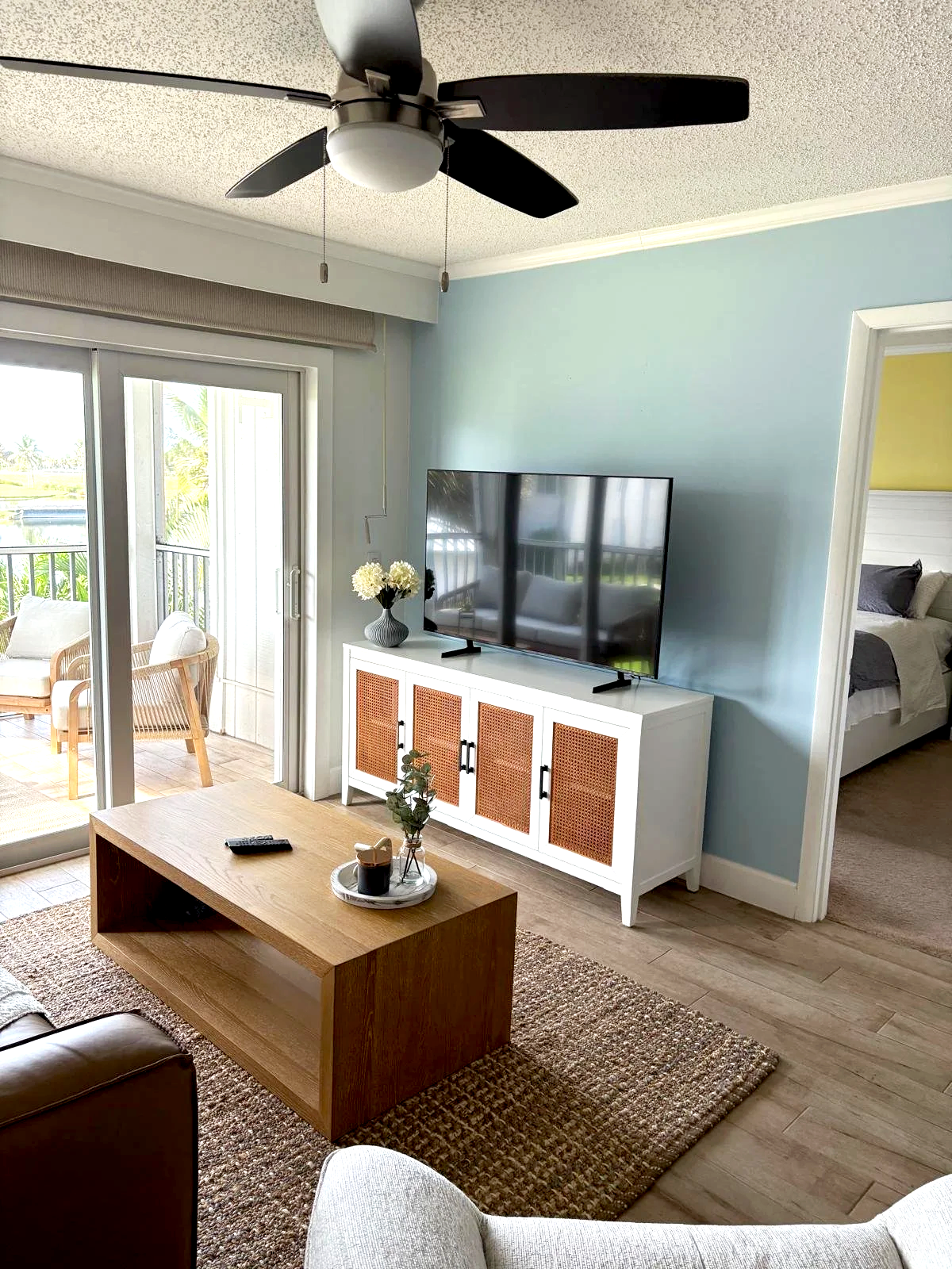 Living room with a sliding glass door leading to a balcony, a wooden coffee table, a white cabinet with rattan panels, a flat-screen TV, a ceiling fan, and a view into a bedroom.