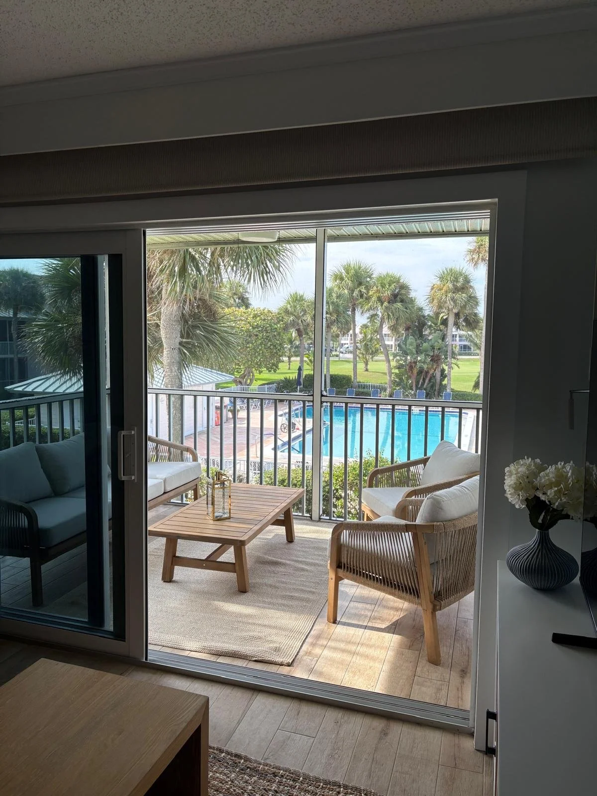 A view from inside a room looking out onto a balcony with outdoor seating, a pool, palm trees, and a grassy area.