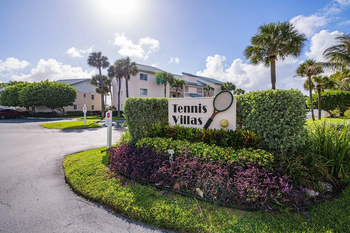 Sign for Tennis Villas with a tennis racket and tennis ball in front of a landscaped area with palm trees and residential buildings in the background.