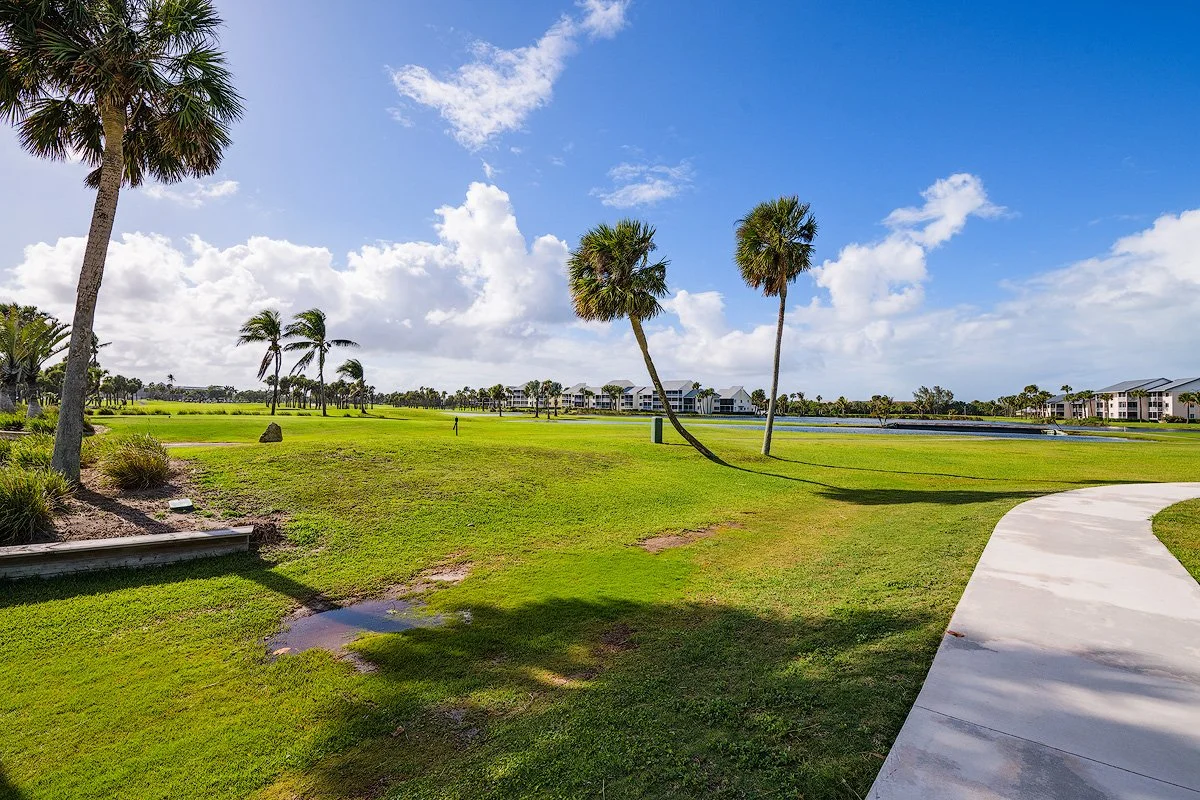 A golf course with several palm trees, a concrete pathway, and a pond surrounded by residential buildings in the distance under a partly cloudy sky.