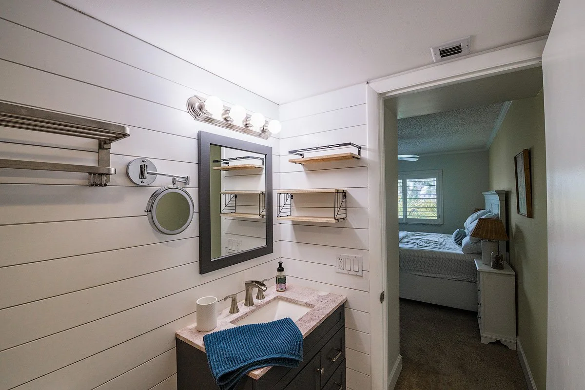 Bathroom with white shiplap walls, a small vanity with a pink countertop and dark cabinet, a mirror, a three-bulb light fixture, wall-mounted shelves, a round wall mirror, a soap dispenser, a blue towel, and a bedroom visible through an open door.