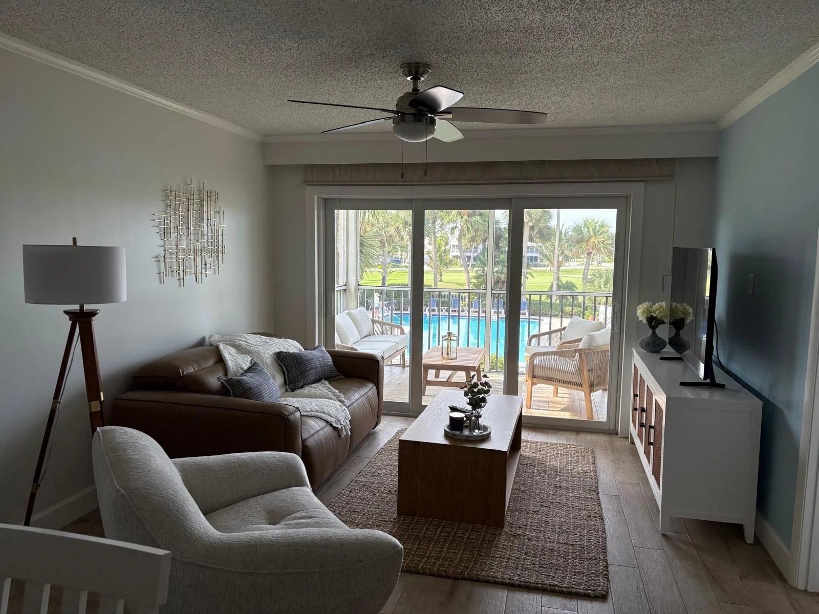 Living room with a sliding glass door leading to a balcony with seating and a view of a swimming pool and palm trees outside.