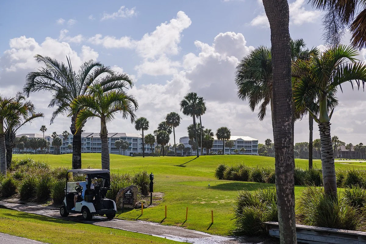 A golf course with palm trees, a golf cart, and a building in the background under a partly cloudy sky.