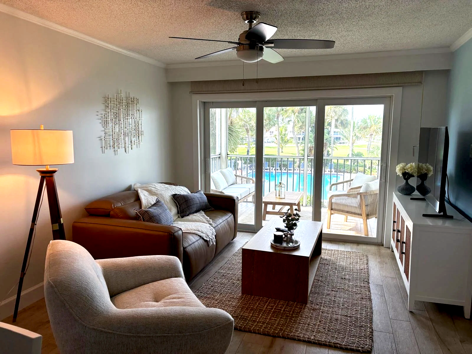 Living room with a sliding glass door leading to a balcony overlooking a pool and palm trees. The room has a beige armchair, a brown leather sofa with throw pillows and a blanket, a wooden coffee table with a small plant, a side table with lanterns, and a white cabinet with vases and a TV. A ceiling fan is above.