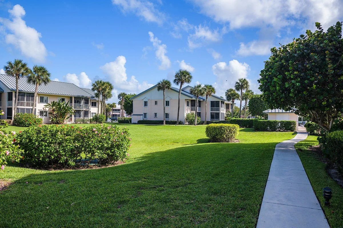 A sunny residential area with well-maintained green lawns, tropical trees, and white buildings with screened porches, under a partly cloudy blue sky.
