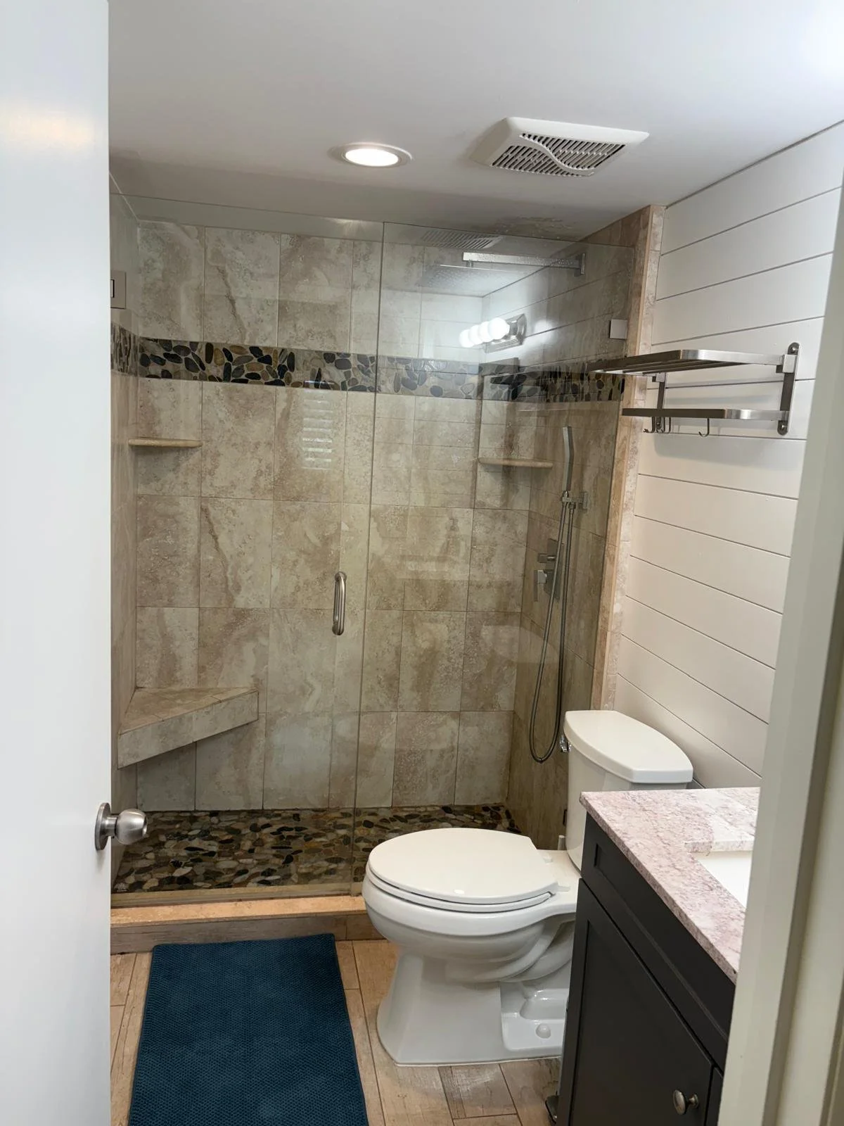 Bathroom with glass-enclosed shower, beige and pebble tile design, white toilet, black vanity, pink granite countertop, and white shiplap wall.