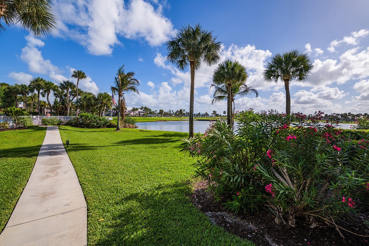 A tropical landscape with a concrete pathway, palm trees, green grass, pink flowers, and a body of water under a partly cloudy sky.