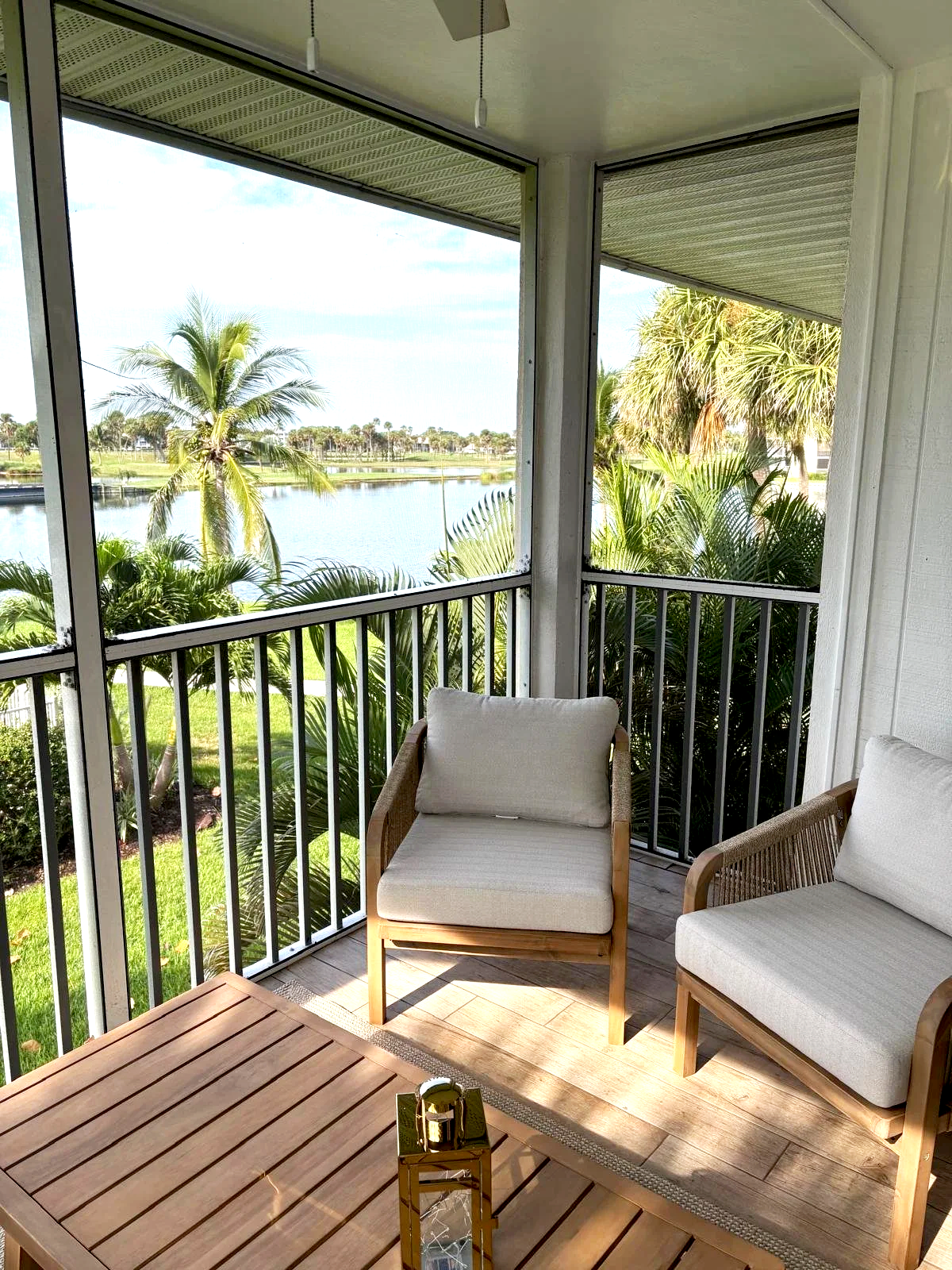 A screened-in balcony with two cushioned chairs around a wooden table, overlooking palm trees and a body of water.