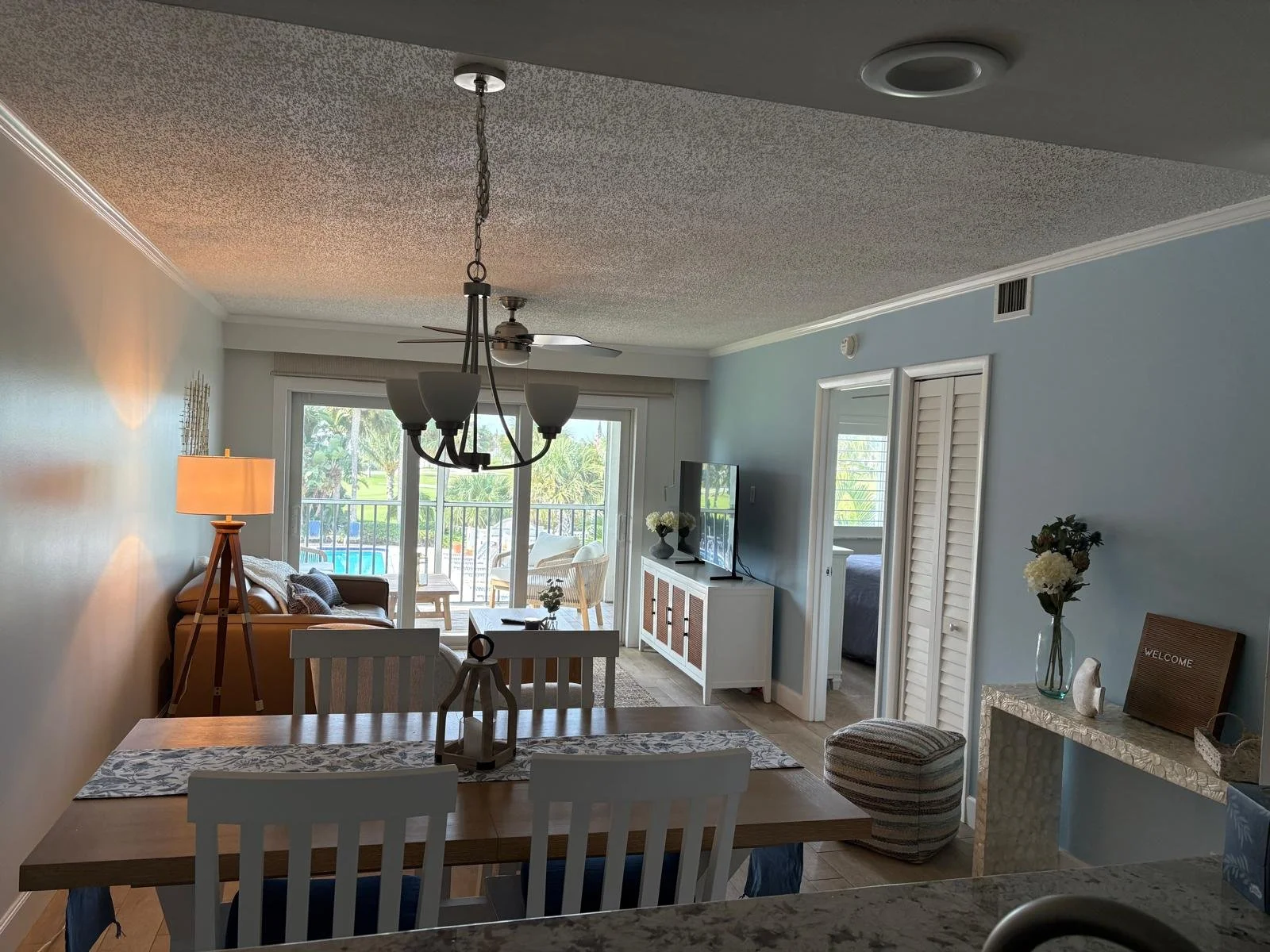 Living room and dining area with sliding glass door to balcony, blue wall, TV, wicker ottoman, and decorative flowers.
