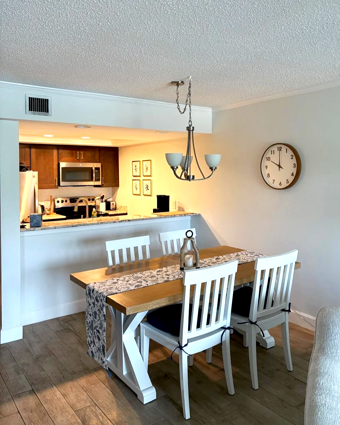 Dining area with wooden table and four white chairs, hanging chandelier, and a wall clock showing 11:53, with a kitchen in the background featuring cabinets, stove, microwave, and refrigerator.