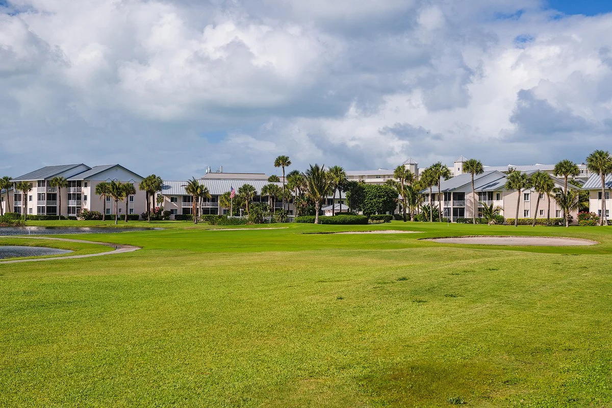 View of a golf course with a sand trap, lush green grass, and palm trees, with residential buildings in the background under a partly cloudy sky.