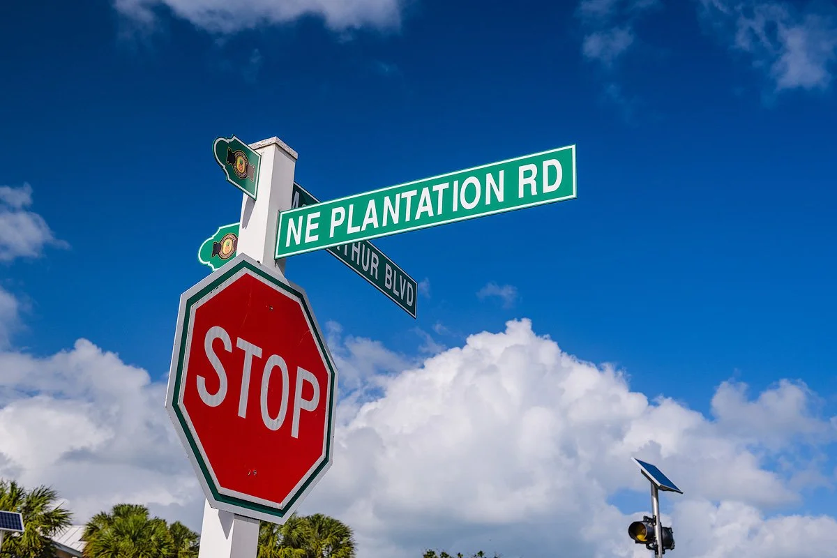 Street signs at the intersection of NE Plantation Road and Thor Boulevard, with a stop sign in the foreground and a partly cloudy blue sky overhead.