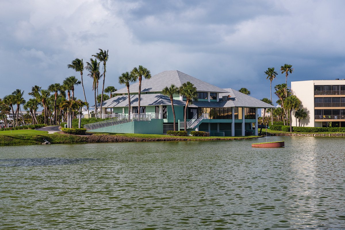 A modern house with a gray roof and pastel green walls located on a small water island with palm trees around it.