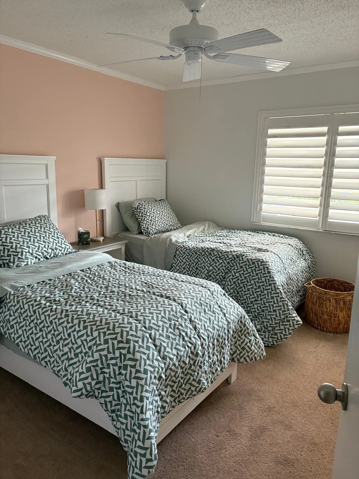 A bedroom with two twin beds featuring matching geometric patterned bedding, a white nightstand with a lamp, and a window with white plantation shutters.