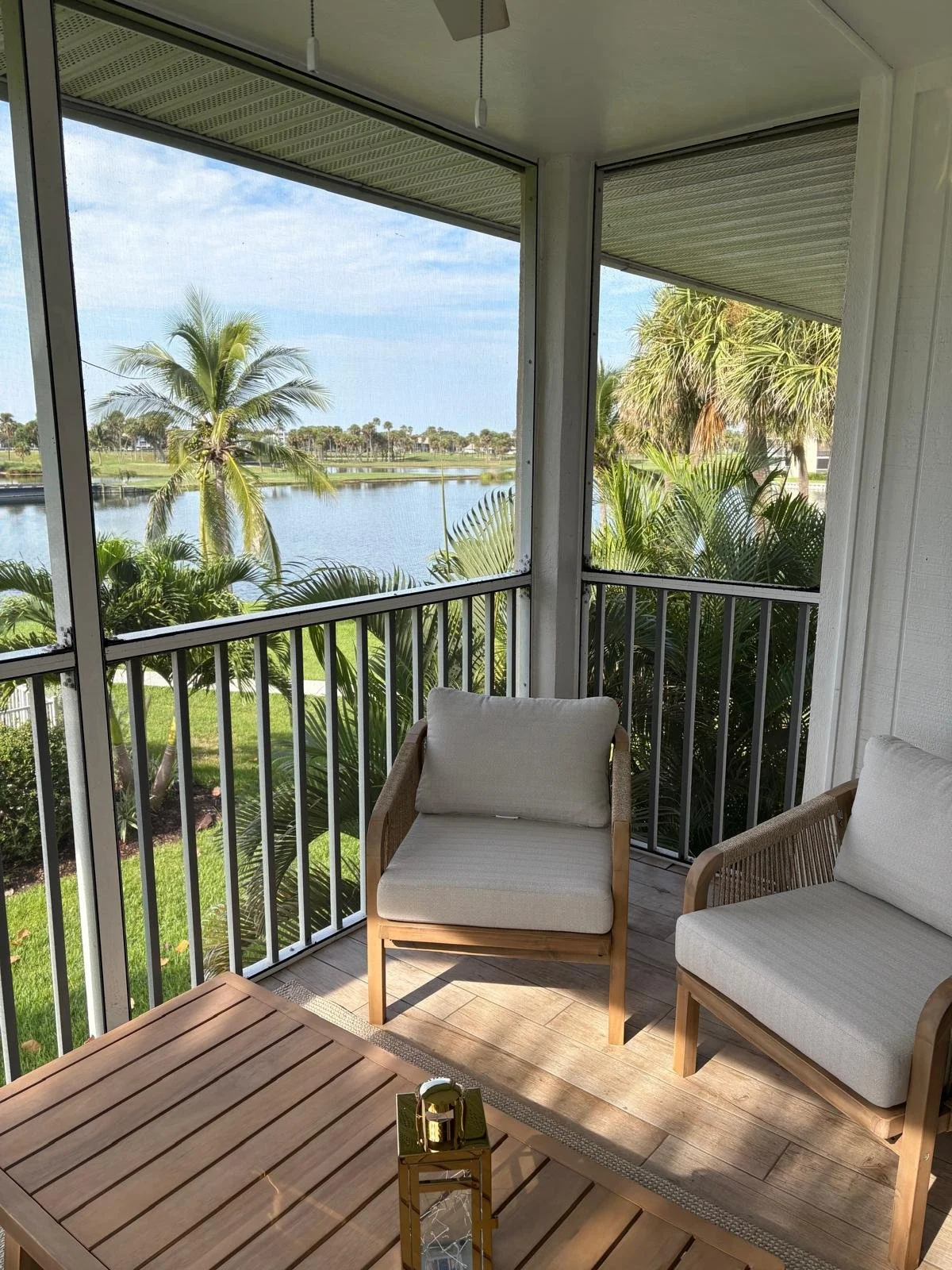 A screened porch with a wooden table and two cushioned chairs overlooking a tropical landscape with palm trees, water, and a blue sky.