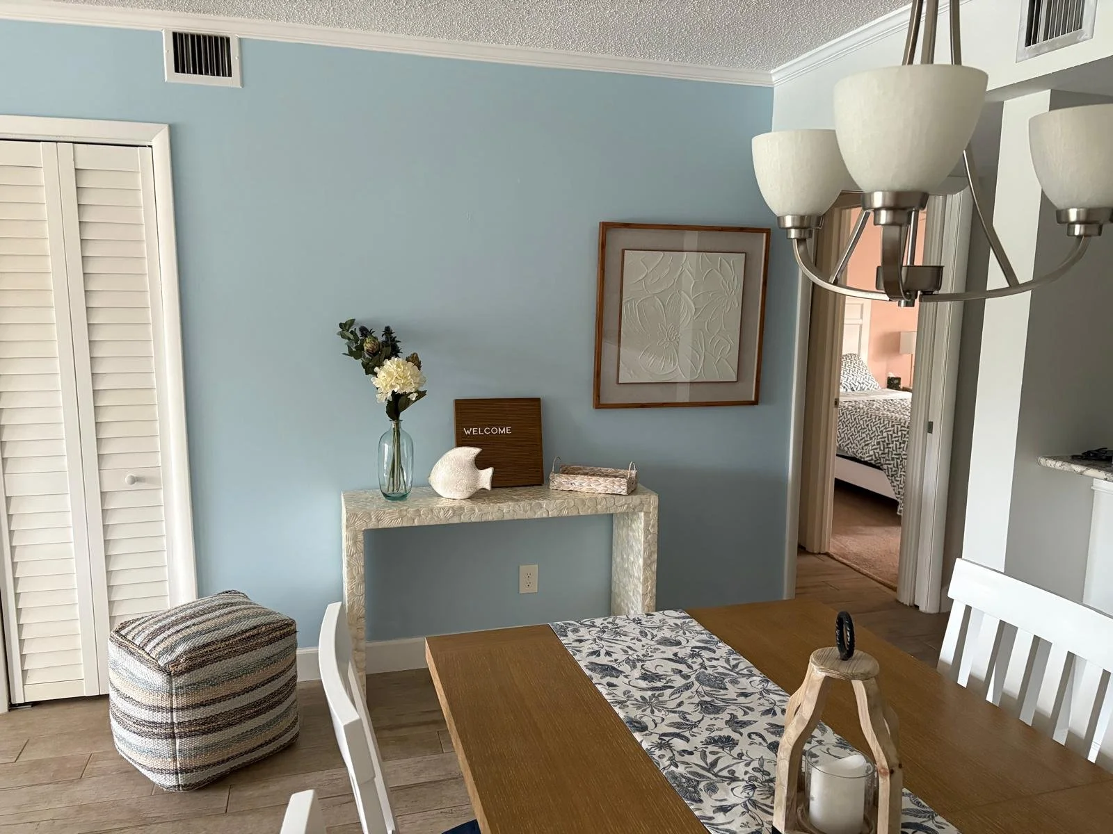 Dining room with a blue wall, a table with a runner, a floral centerpiece, and a chandelier. A doorway reveals a bedroom with a bed and patterned bedding.
