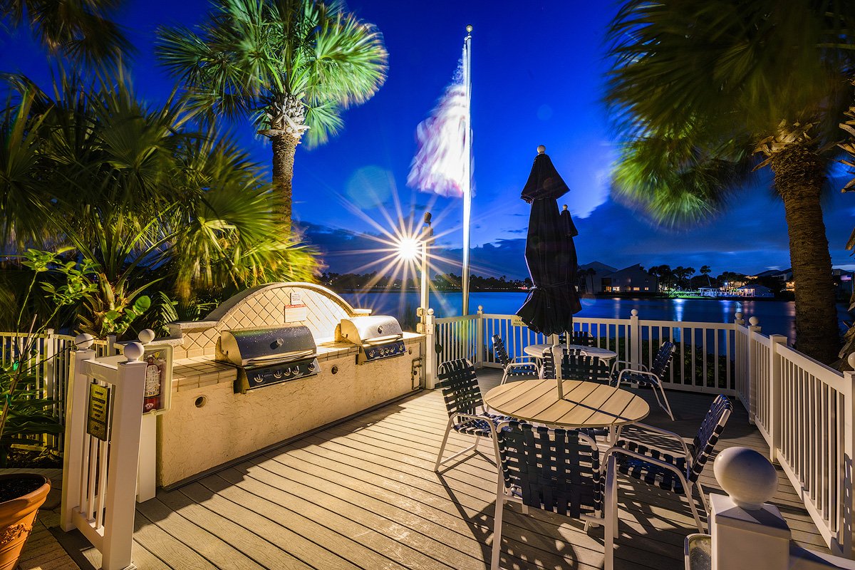 Balcony or patio area with outdoor grill, tables, and chairs at twilight, overlooking a body of water with trees and houses in the distance, surrounded by tall palm trees.