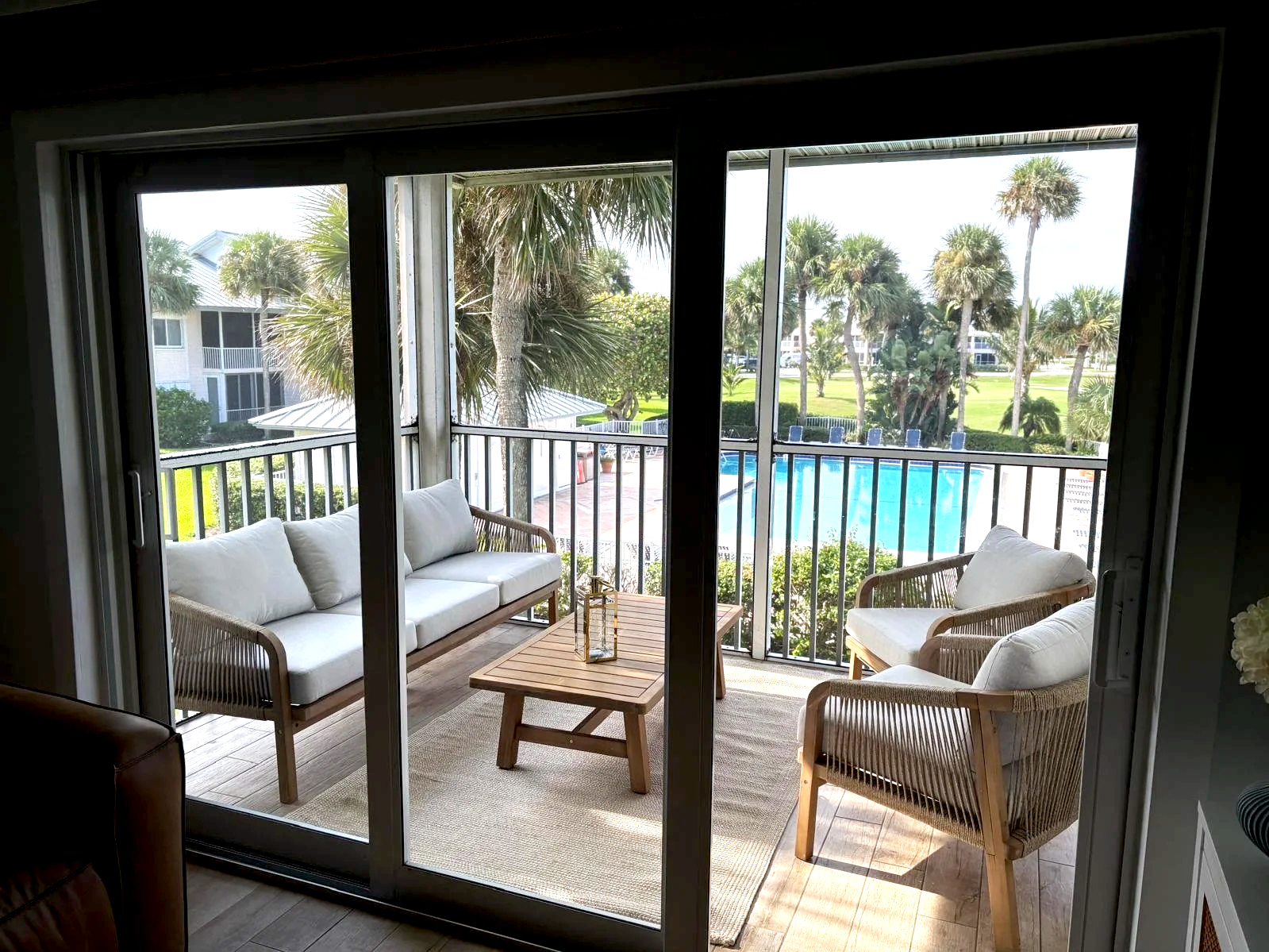 View of a screened balcony with white cushioned outdoor furniture, a wooden coffee table, overlooking a swimming pool with palm trees and a grassy area.