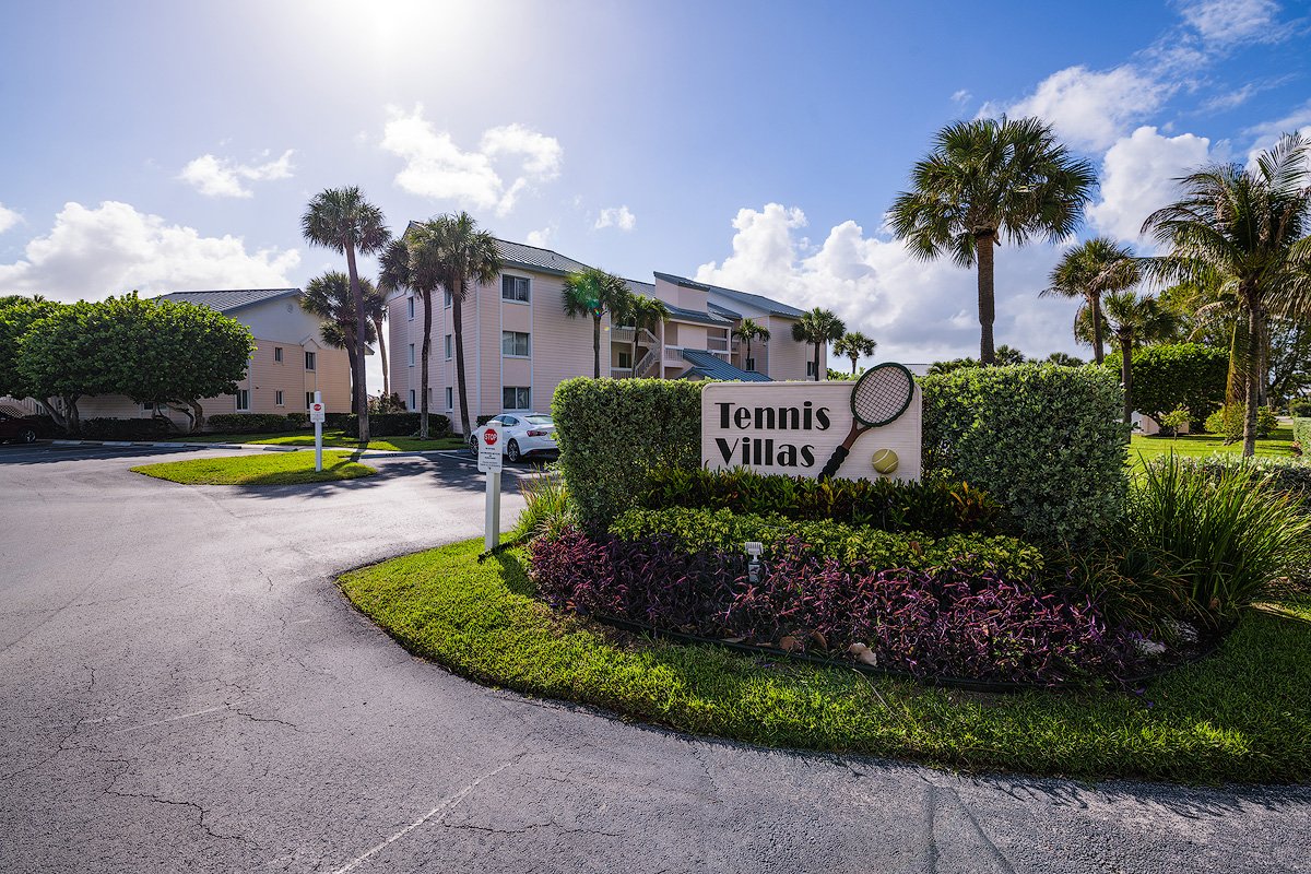 Entrance to Tennis Villas with a sign featuring a tennis racket and ball, surrounded by green bushes and palm trees, with a multi-story apartment building in the background under a partly cloudy sky.