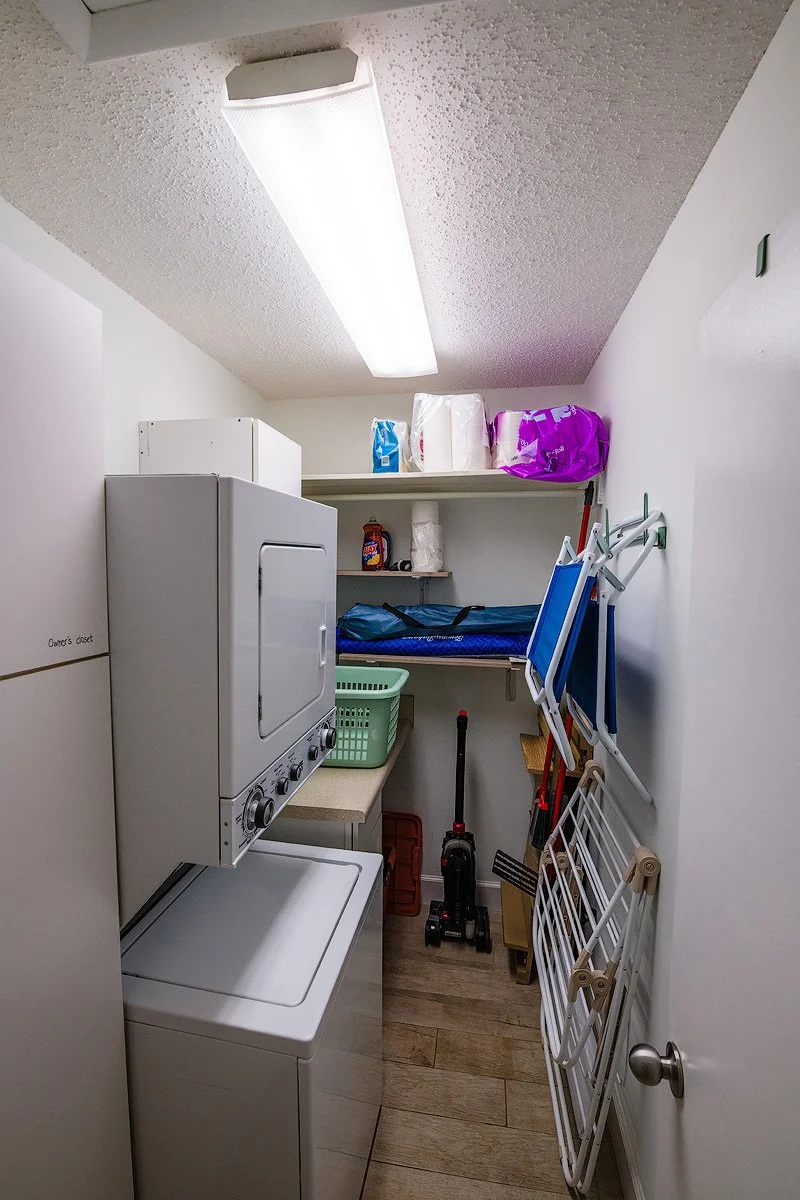 Small laundry room with a stacked washer and dryer, shelves with laundry supplies, an ironing board, cleaning tools, and a small basket.