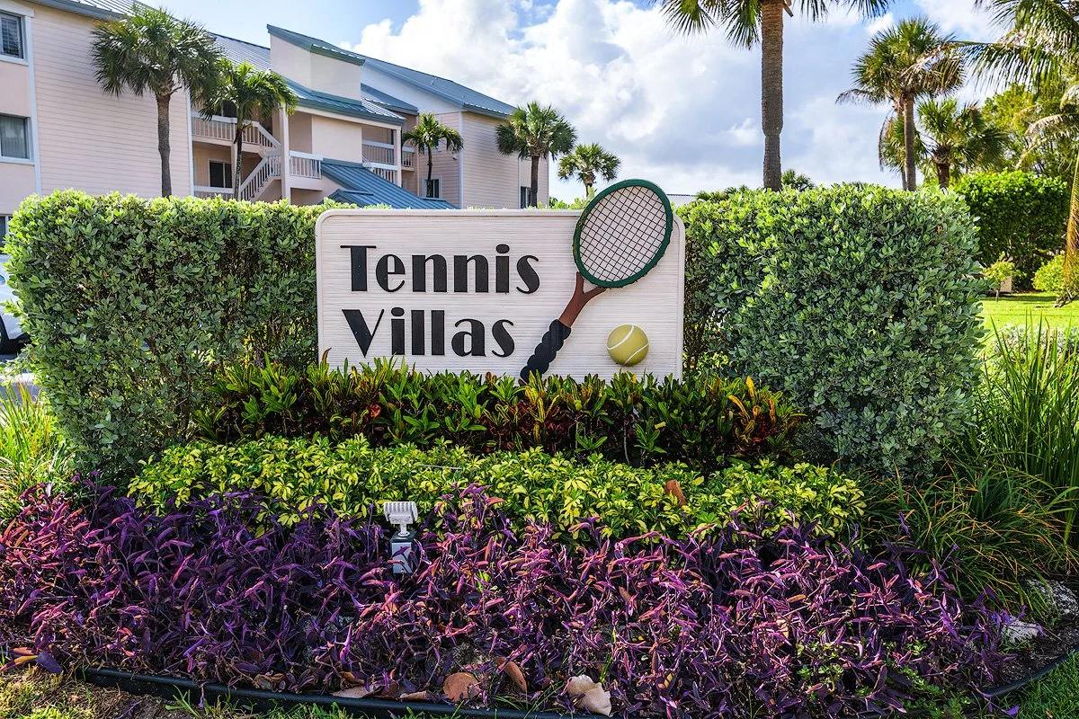 A sign that says "Tennis Villas" with a tennis racket and ball graphic, surrounded by lush green bushes, purple plants, and palm trees in the background.