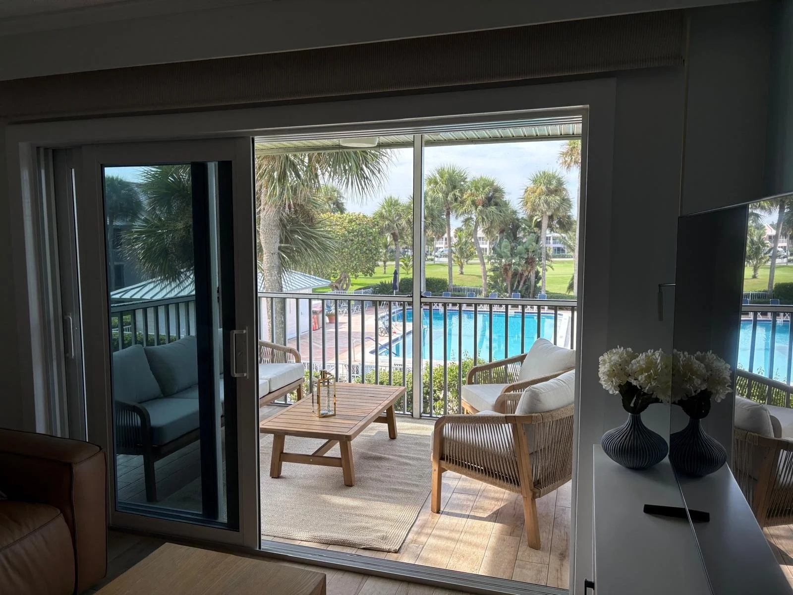 View from inside a living room looking out onto a balcony with outdoor furniture, overlooking a pool area with palm trees and green landscape.