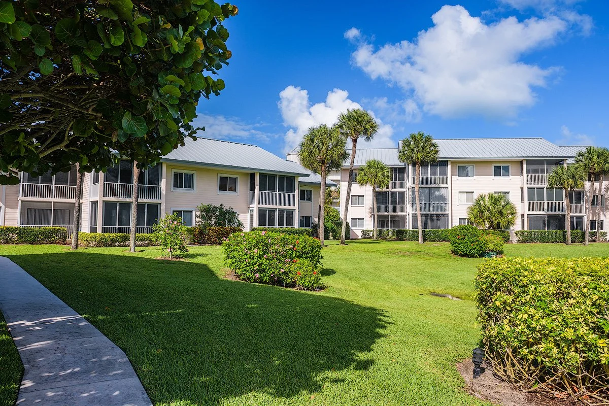A landscaped area with green grass, bushes, and trees in front of a beige apartment building with balconies, under a partly cloudy blue sky.
