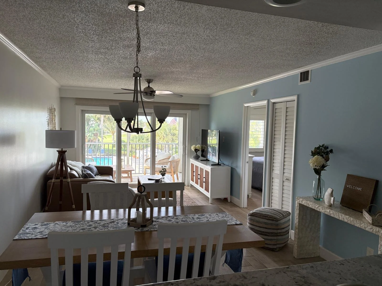 Living room with sliding glass door leading to outdoor patio, dining table with white chairs, sofa with pillows, TV on white cabinet, seafoam wall, and sliding closet doors.