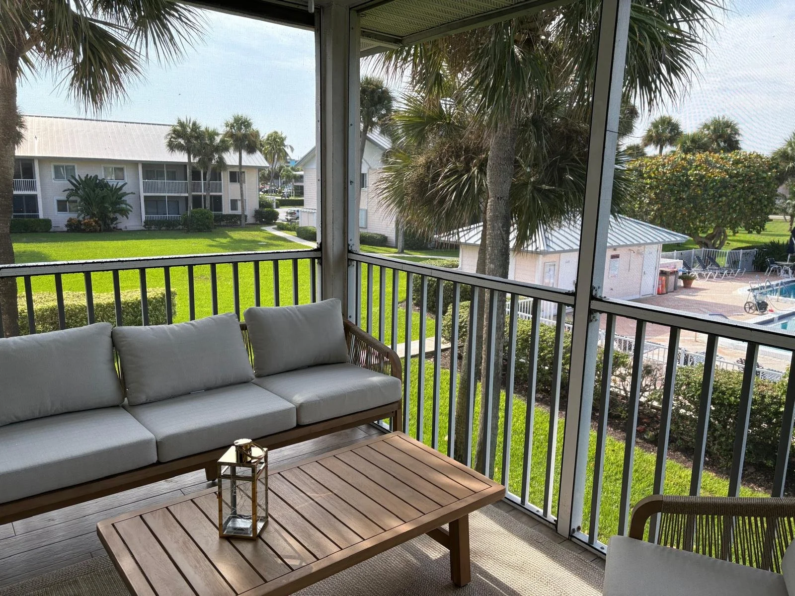 View of an outdoor balcony with a wooden table, a lantern, and a cushioned sofa, overlooking a landscaped area with palm trees, grass, and nearby buildings and a pool.
