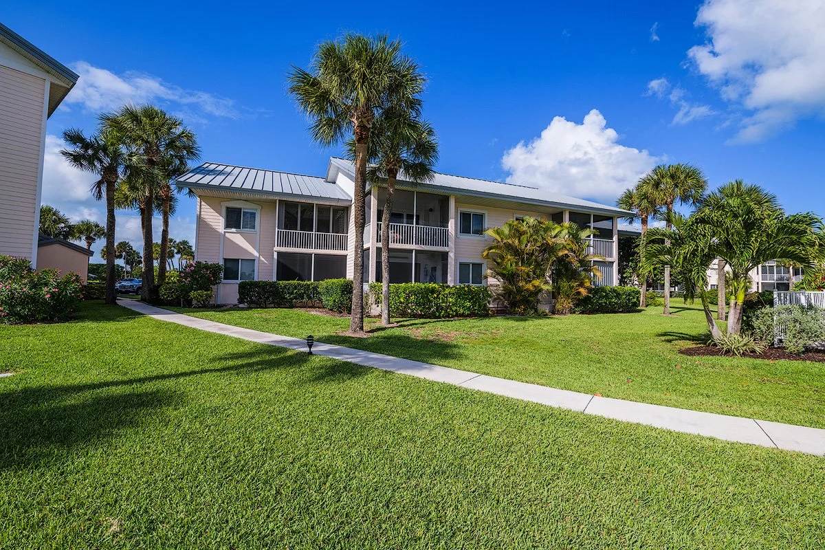 Two-story apartment building with screened balconies, surrounded by well-maintained grass, palm trees, and shrubs, under a partly cloudy sky.