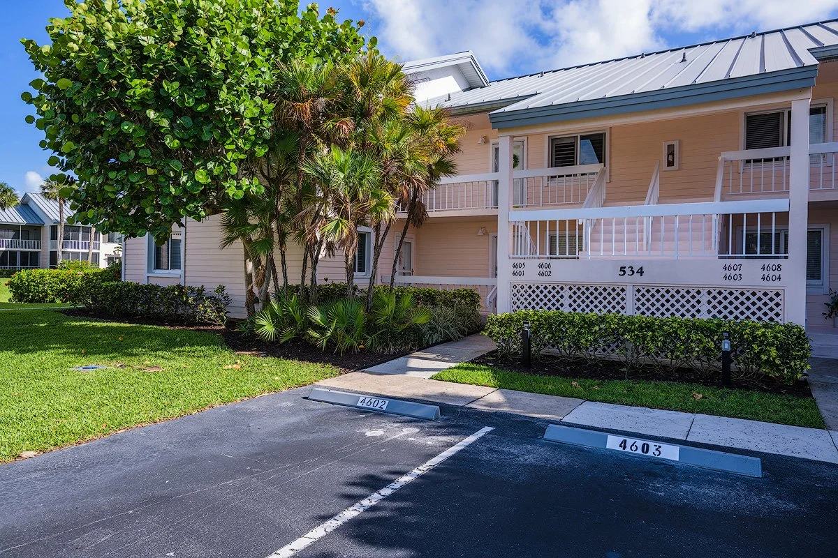 Exterior view of a two-story residential building with beige siding, white railings, and a metal roof. There are parking spots in the foreground numbered 4602 and 4603, with well-maintained landscaping and palm trees surrounding the building.