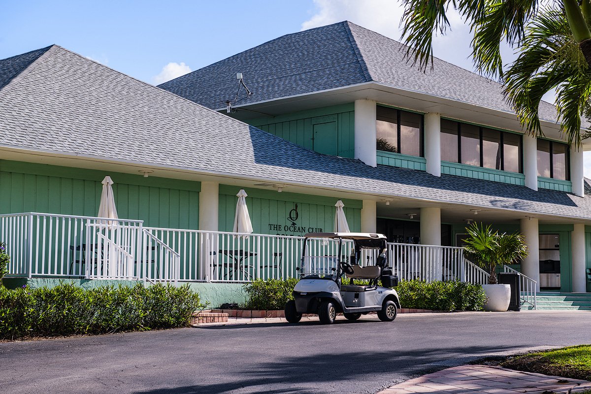 A two-story building with a sign that reads "The Ocean Club" on the front. The building has pale green walls, large windows on the upper floor, and a roof with gray shingles. There is a white railing surrounding a patio area with umbrellas and chairs