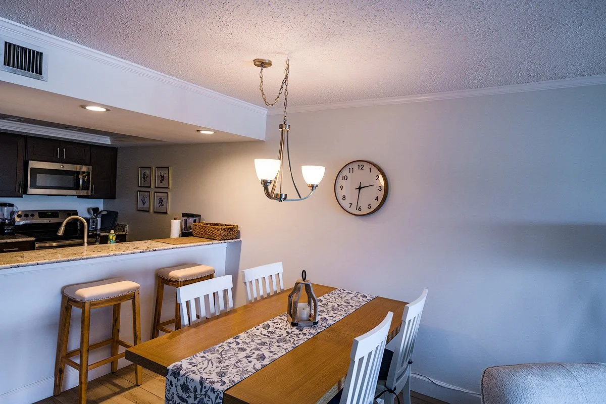 Dining area with a wooden table, white chairs, and a decorative runner. Overhead light fixture with three bulbs. Kitchen in the background with dark cabinets, microwave, and stovetop. Wall clock showing approximately 2:01.