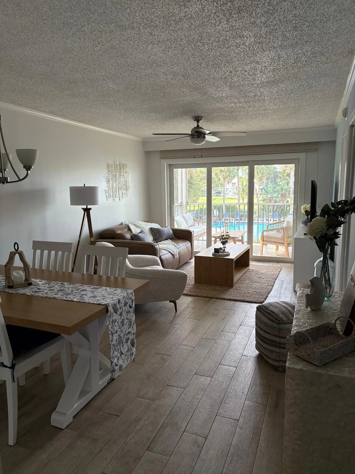 Living room with beige couches, wooden coffee table, sliding glass door leading to balcony, ceiling fan, area rug, TV, floral arrangements, and wooden flooring.