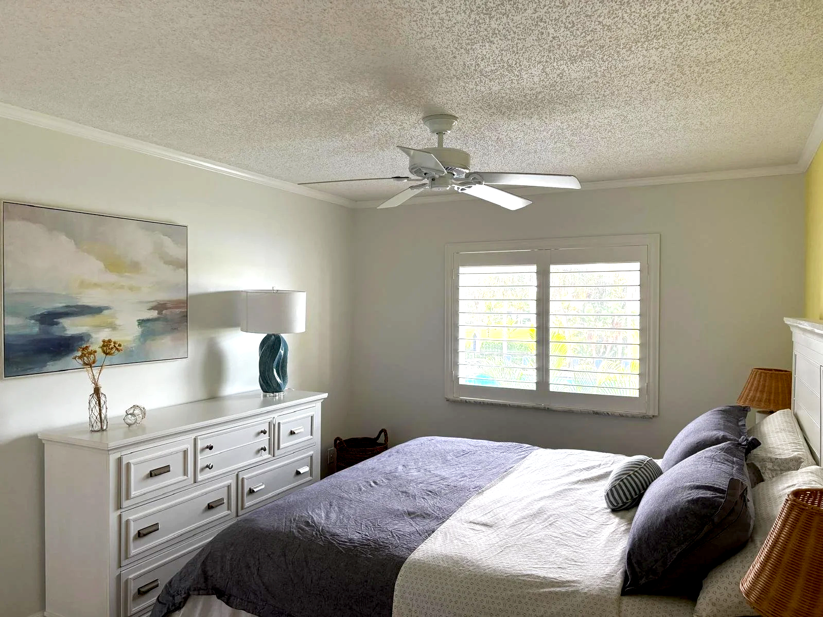 A bedroom with a white dresser, a table lamp, and framed painting. The bed has dark pillows and a wicker lamp on the side. A window with white shutters is in the background.