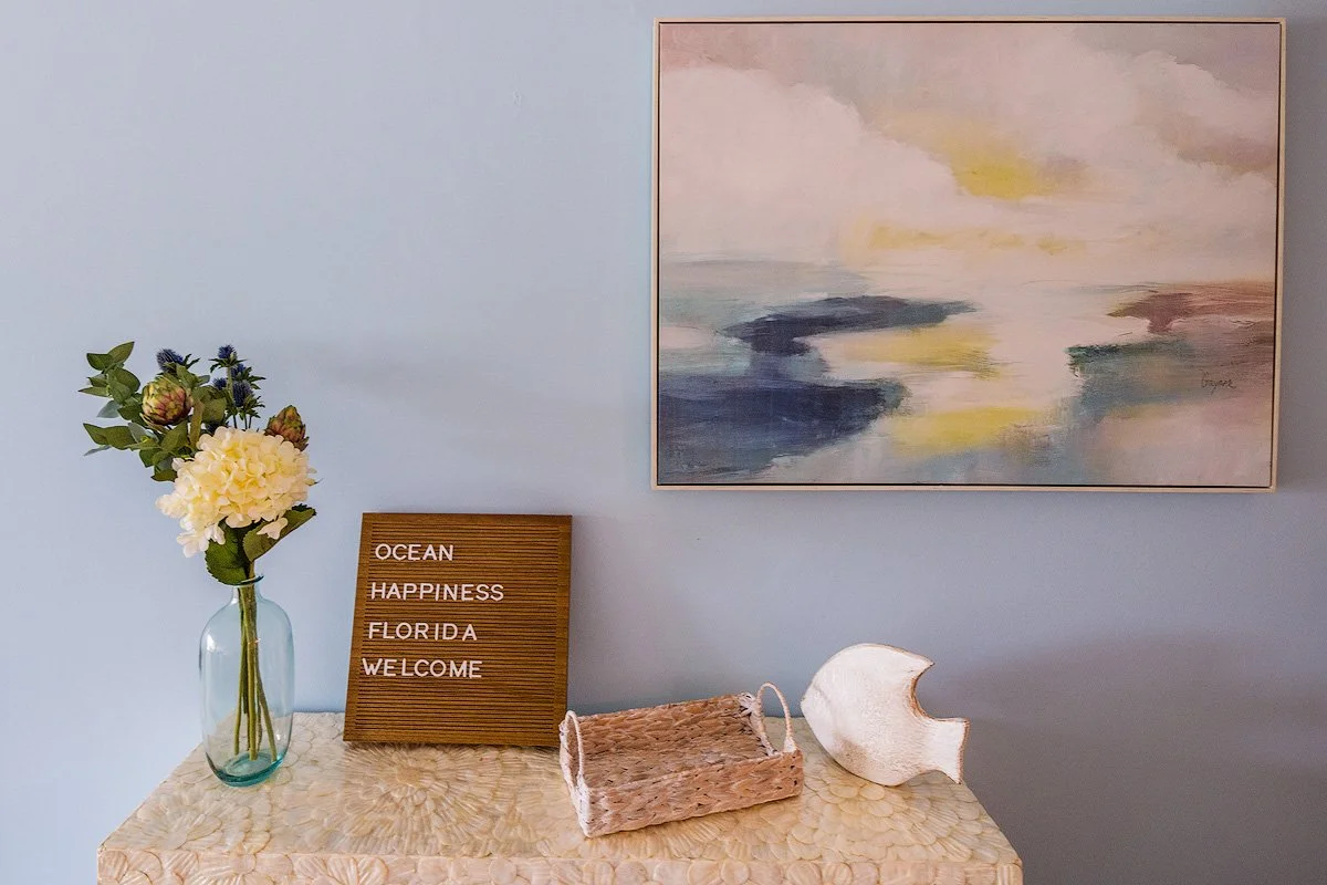 Decorative coastal-themed interior scene with a vase of flowers, a wooden letter board with "OCEAN HAPPINESS FLORIDA WELCOME," a woven basket, and a large white seashell on a textured surface against a light blue wall.