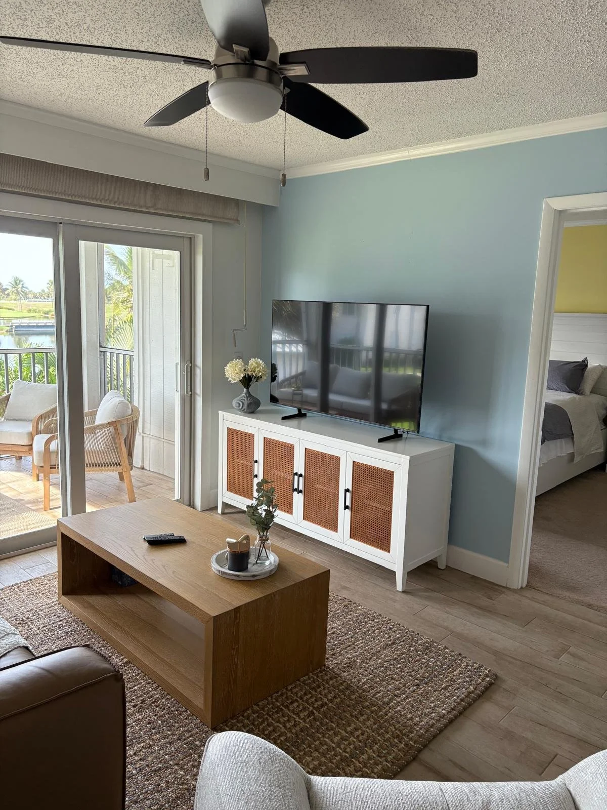 Living room with light blue walls, a flat-screen TV on a white cabinet, a wooden coffee table with a remote and decorative items, a sliding glass door leading to a balcony with outdoor furniture, a ceiling fan, and a view of a tropical landscape outs