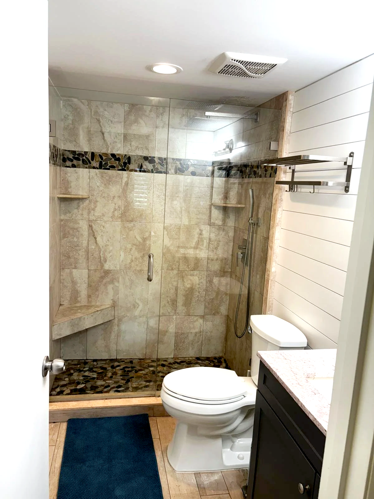 Bathroom with a glass-enclosed shower area, a toilet, and a dark vanity with a pink countertop. The shower has beige tiles with a decorative pebble border and built-in corner shelves. A blue bath mat is on the wooden floor in front of the toilet.