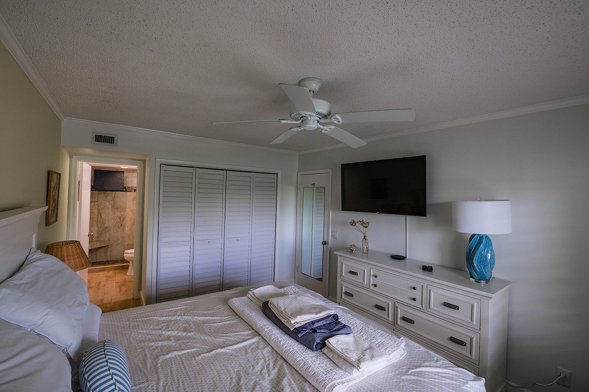 Well-lit bedroom with a bed, dresser, wall-mounted TV, ceiling fan, and bathroom visible through an open door.