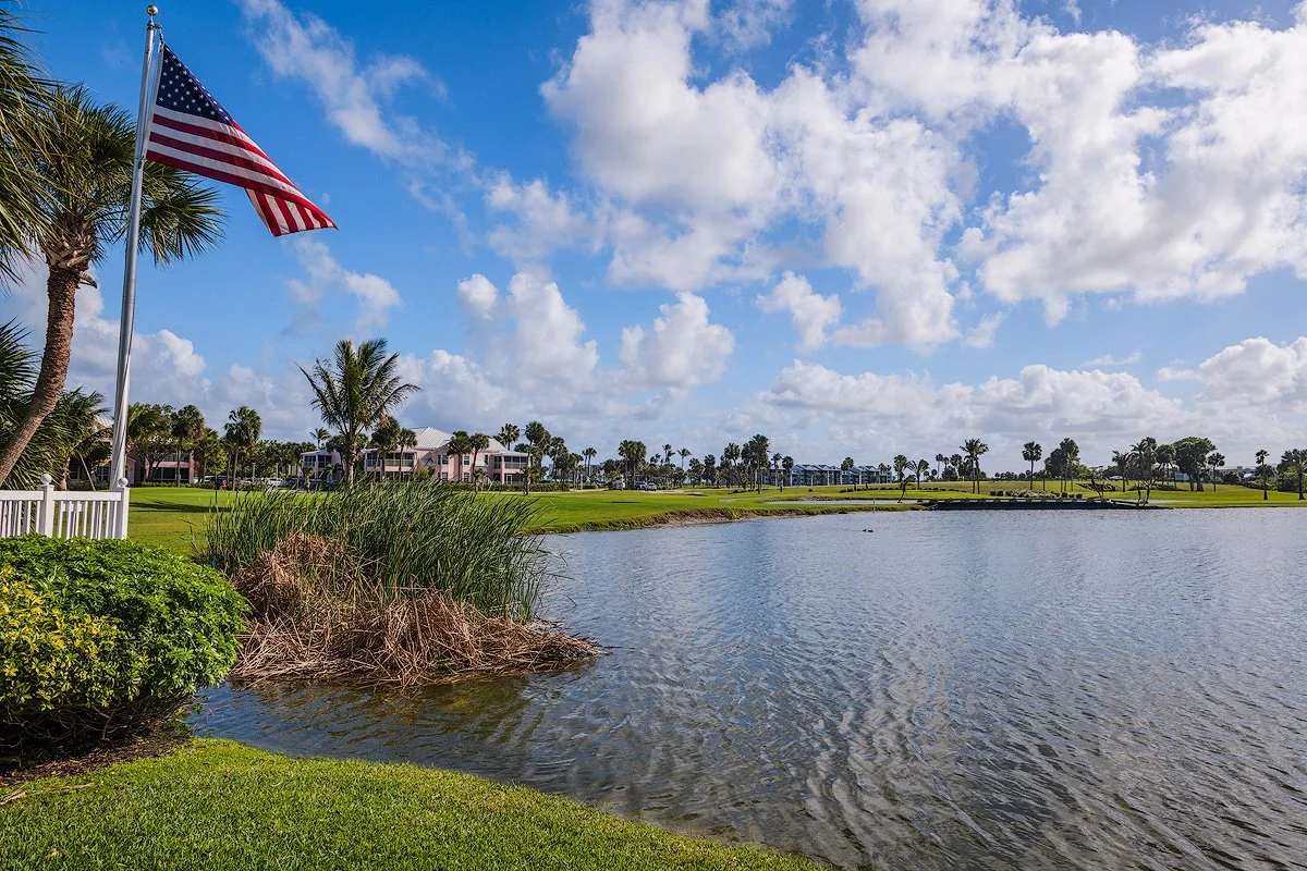 A scenic view of a park near a body of water with an American flag on a flagpole, palm trees, green grass, residential buildings in the background, and a partly cloudy sky.