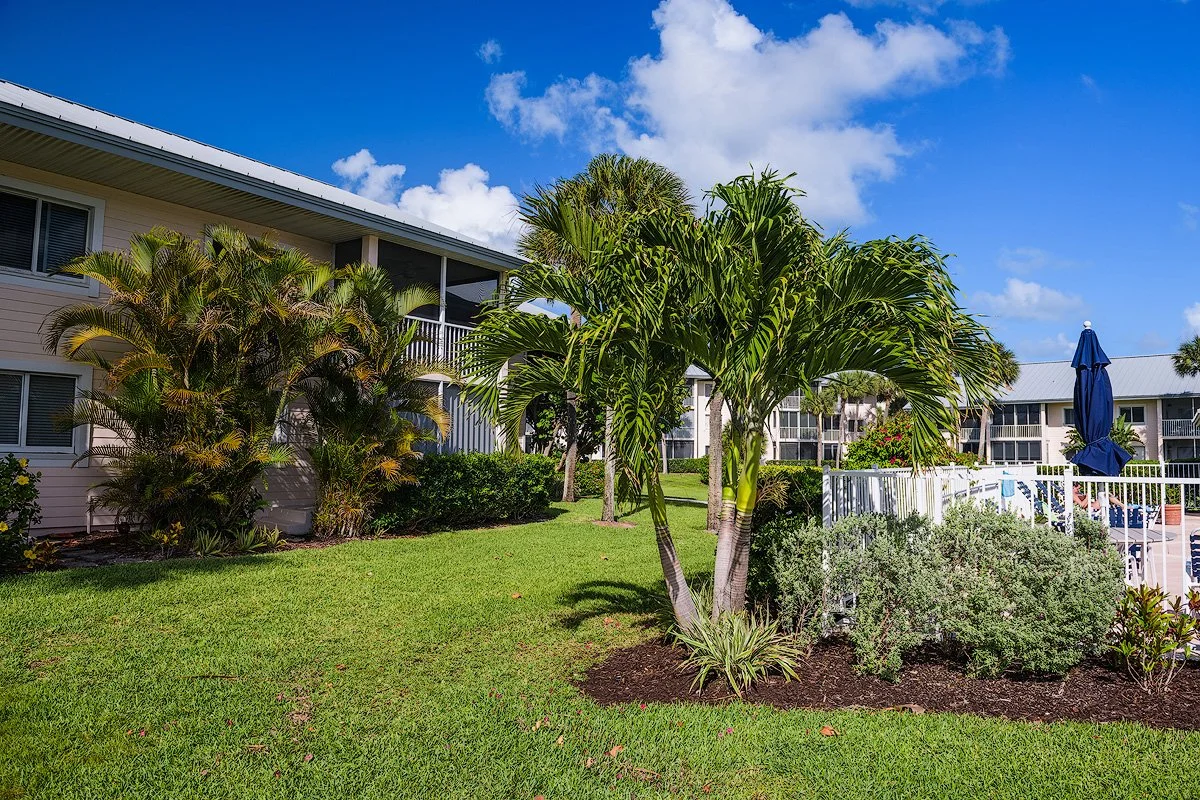 A tropical garden with palm trees, green grass, and bushes in front of a white building with balconies under a blue sky with some clouds.