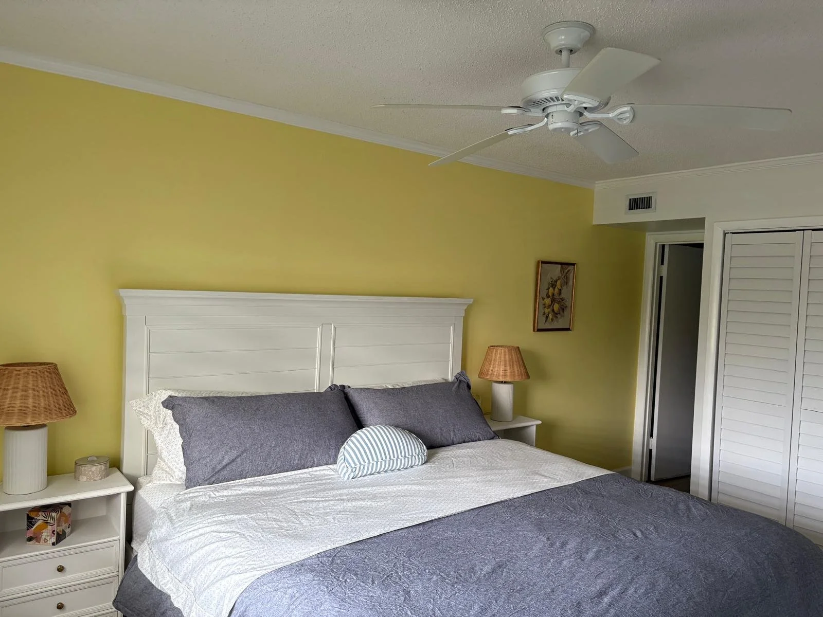 A bedroom with a white headboard, grey pillows and a white and grey bedspread, yellow accent wall, two wicker lamps on white nightstands, a ceiling fan, a framed picture on the wall, and white louvered closet doors.