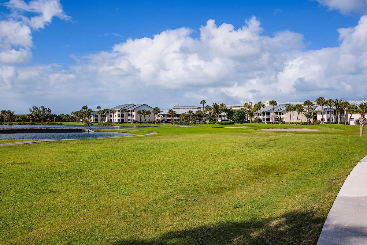View of a golf course with green grass, a pond, palm trees, and residential buildings in the background under a partly cloudy sky.