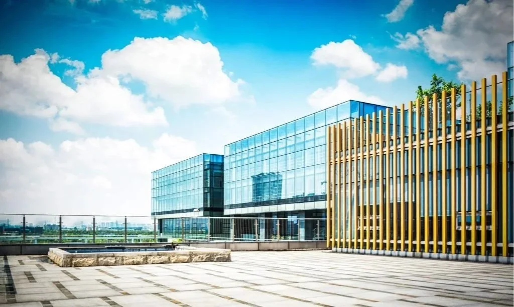 Modern glass building with a spacious rooftop terrace, featuring glass walls, wooden slats, and a city skyline in the background under a partly cloudy sky.