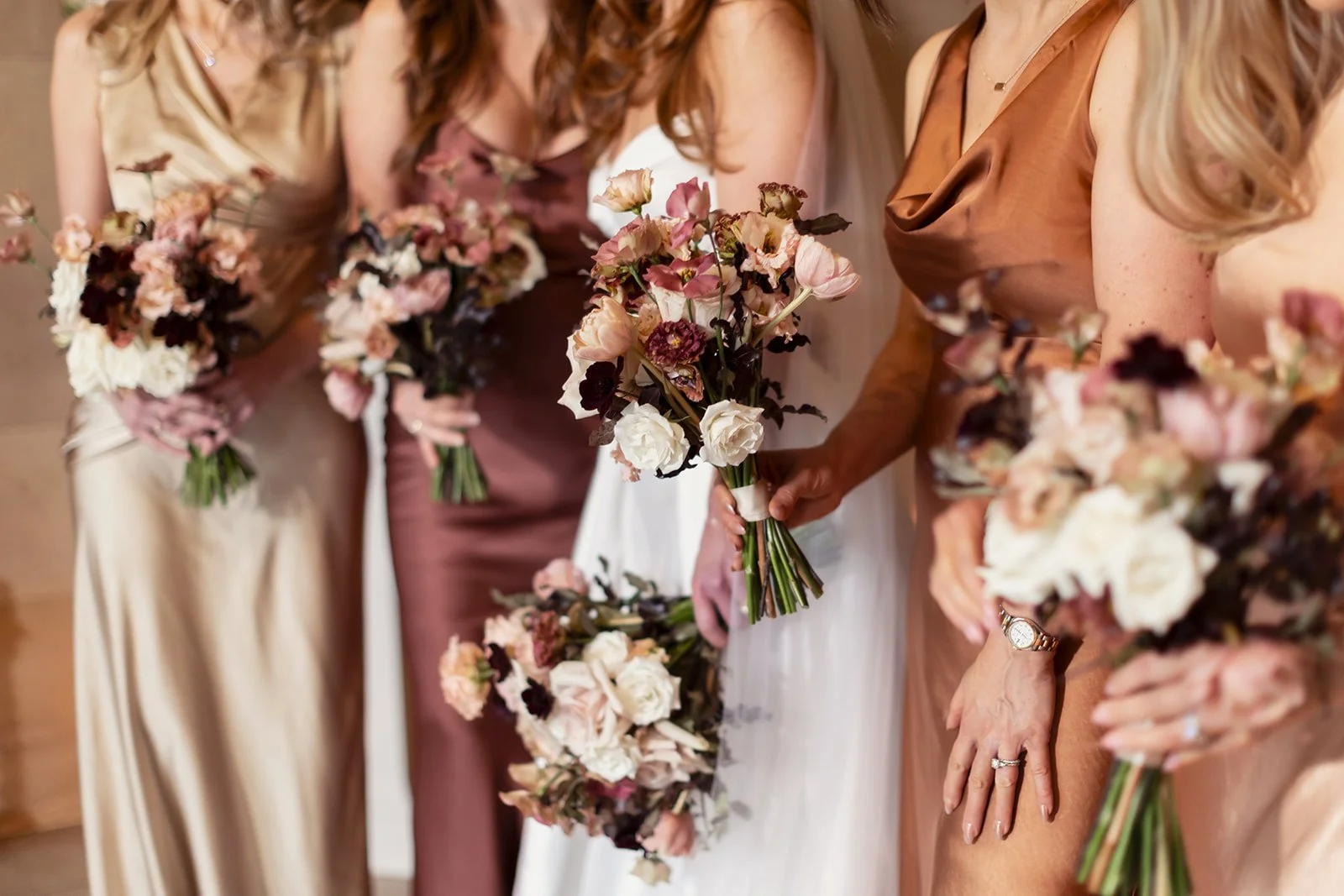 Close-up of women, including a bride and bridesmaid, dressed in elegant attire with autumnal tones, holding bouquets of flowers at a wedding.