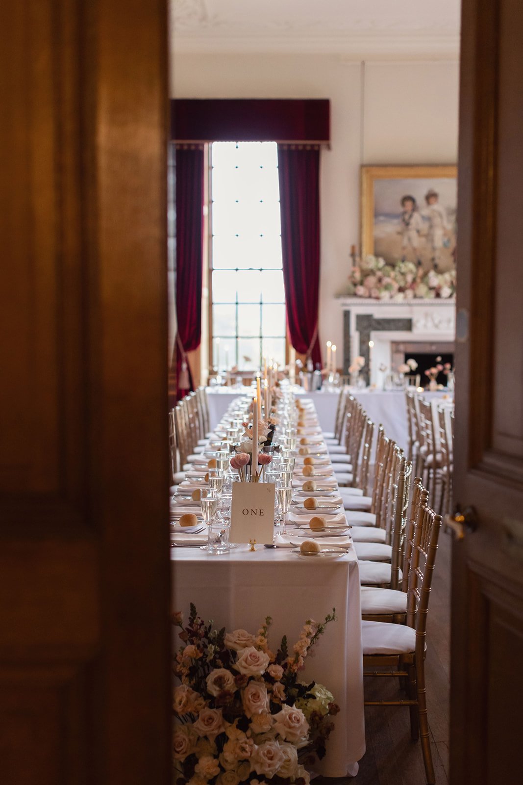 Elegant dining room in a Scottish castle, with a long table set for a wedding, decorated with flowers, candles, and name cards, viewed through a partially opened wooden door.