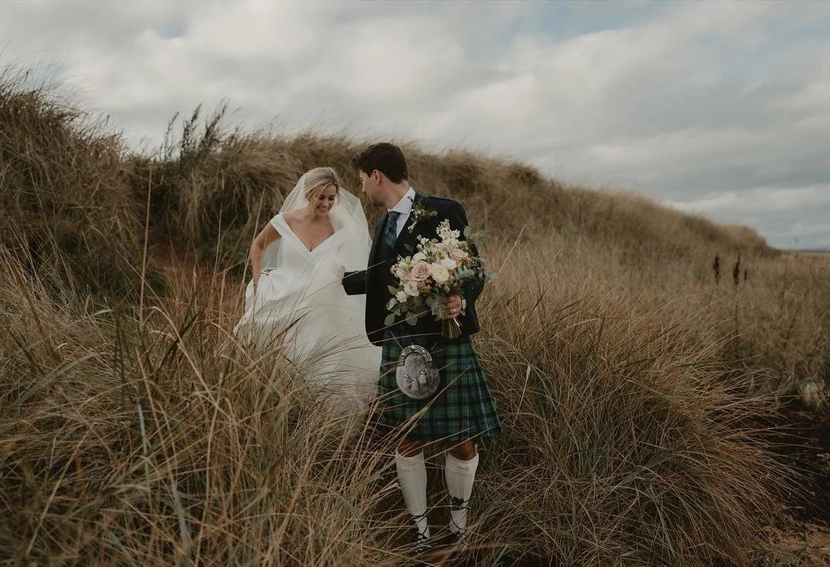 A bride in a white wedding dress and veil, and a groom in a kilt, standing together in tall grass on a hilltop, with a cloudy sky overhead, during a wedding photoshoot.
