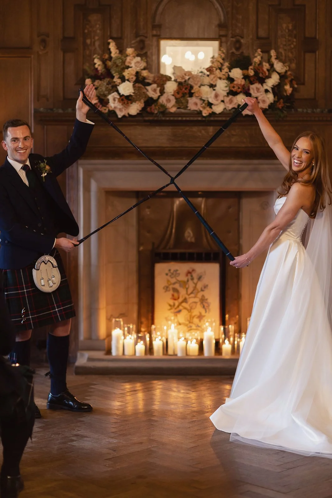 A bride and groom in wedding attire holding tartan handfasting ribbons that signify and symbolise their marriage, smiling at each other and their wedding guests in a large room with a fireplace, flowers, and candles.