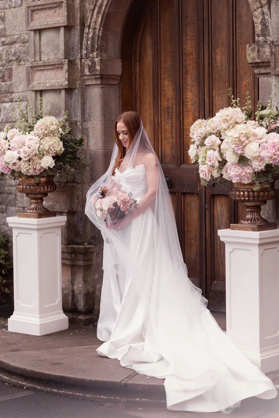 A bride in a white wedding gown standing outside a stone built Scottish castle with a wooden door, holding a bouquet of soft pink and white flowers, with large floral urn arrangements on white pedestals on either side.