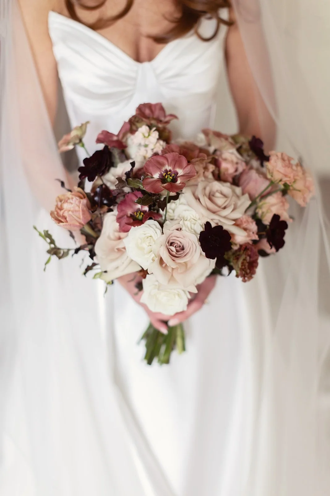 Close-up of a bride holding a bouquet of soft pink, natural whites, and pops of dark-colored burgundy flowers, wearing a white wedding dress with veil.